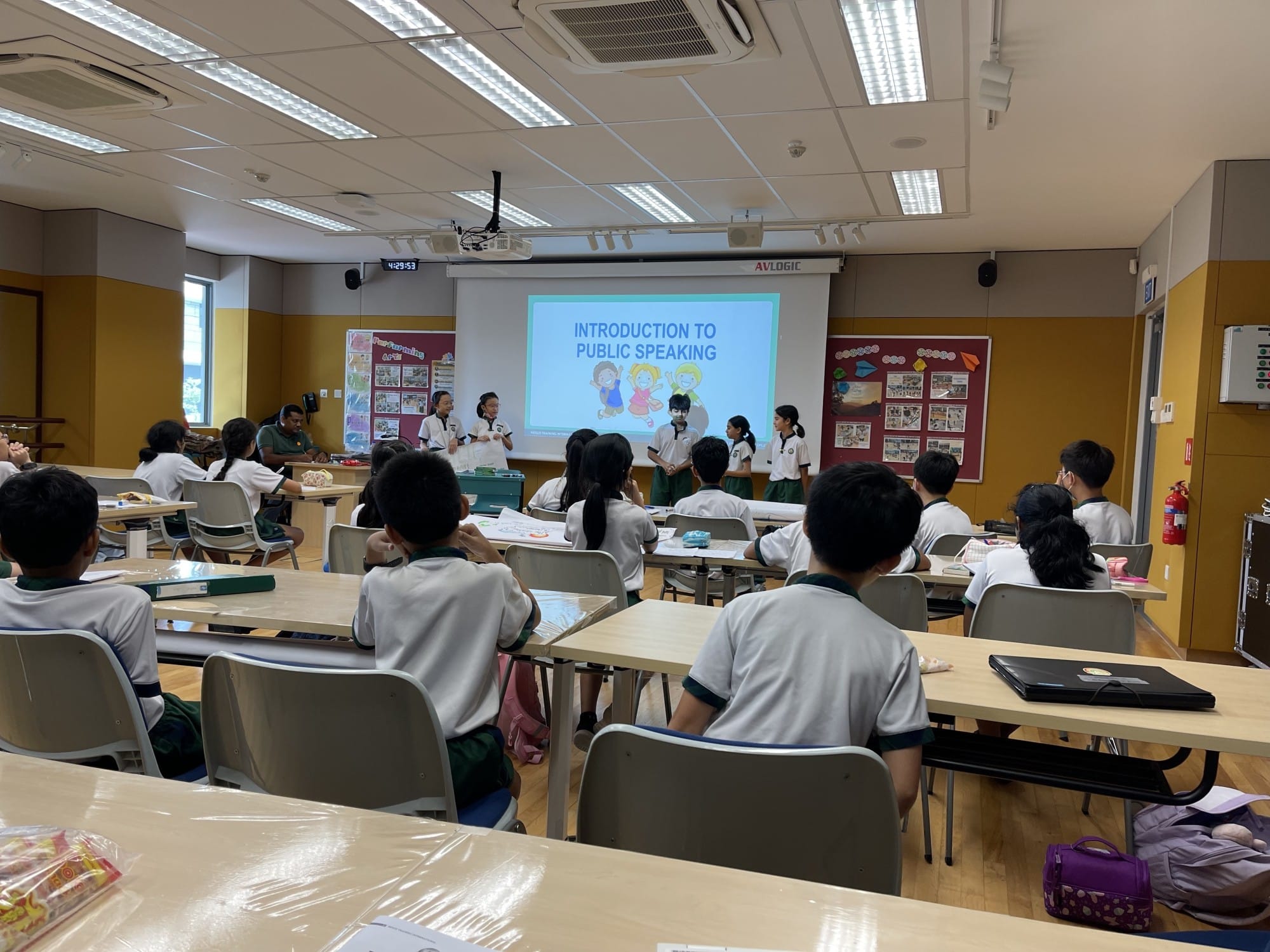 Students in uniforms sit at desks facing peers presenting in front of a projected "Introduction to Public Speaking" slide.